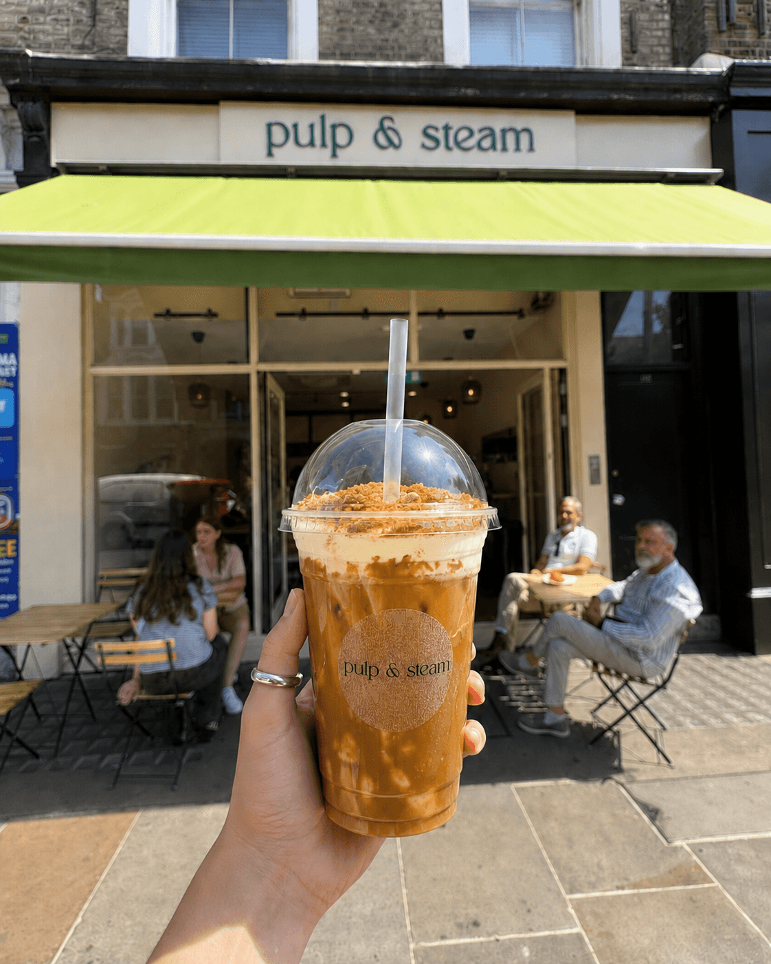 An iced coffee in a Pulp & Steam cup held up in front of the shop on Ladbroke Grove